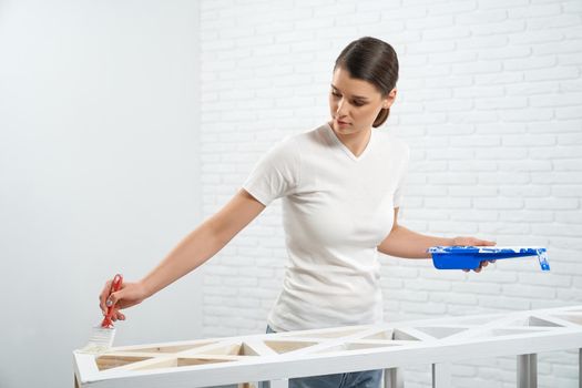 Beautiful young brunette in white shirt painting wooden rack in empty room. Concept of repair at home.