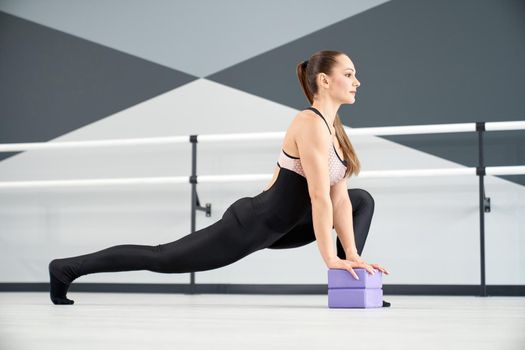 Side view of stunning female dancer practicing lunges in dance hall. Gorgeous fit woman wearing black tight clothes stretching legs using small purple blocks on floor. Stretching, gymnastics concept.