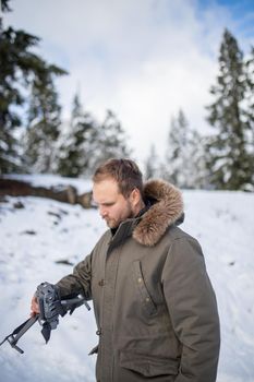 Handsome man holding drone with snowy pines as background. Portrait of angry-looking man interacting with drone in frozen woods. Adventurous winter holiday