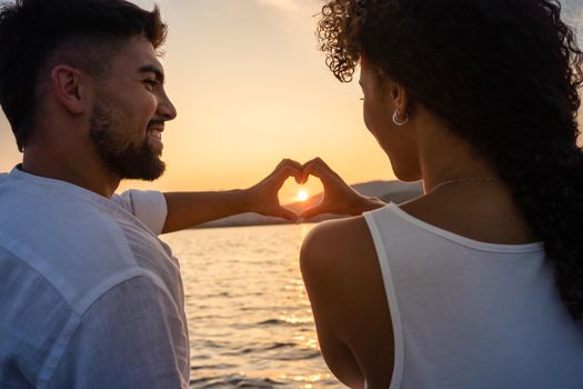Young just married multiracial couple in love making heart shape with hands with setting sun in centre of it, looking in the eyes smiling. Romantic scene of Hispanic girl and boyfriend view from back