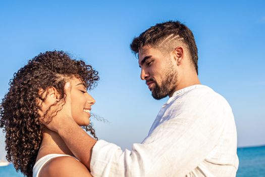 Moment of tenderness between young multiracial couple of happy lovers who look into each other's eyes while he smilingly holds her head in hands. Passion and love of two Latin people on sea vacations