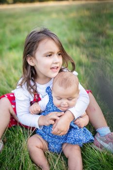 Adorable little girl sitting on grass and lovingly hugging baby. Portrait of cute young child sitting with her baby sibling in park. Young family playing outdoors