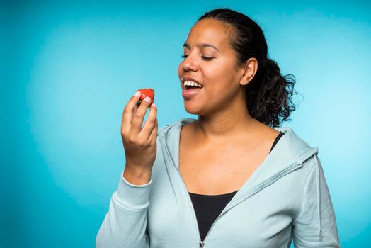 Beautiful young mixed race woman in casual clothing eating and enjoying a fresh strawberry with a blue background