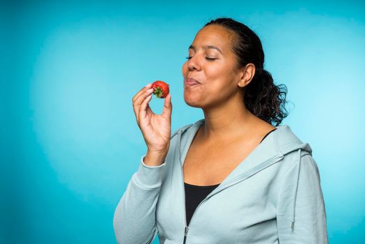 Beautiful young mixed race woman in casual clothing eating and enjoying a fresh strawberry with a blue background