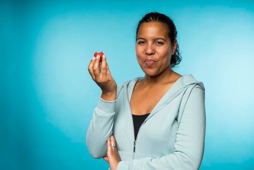 Beautiful young mixed race woman in casual clothing eating and enjoying a fresh strawberry with a blue background