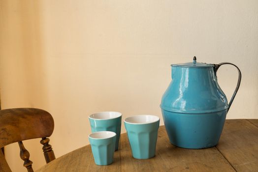 Blue jar with three blue cups on a wooden table with a chair indoors