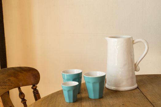 white jar or jug with blue cups on a wooden table with a chair