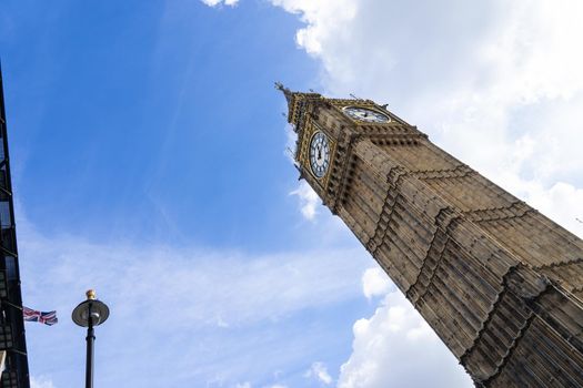 Low angle view of the Big Ben in London England United Kingdom against a blue sky with white clouds and a Union Jack British flag on a building and a streetlight