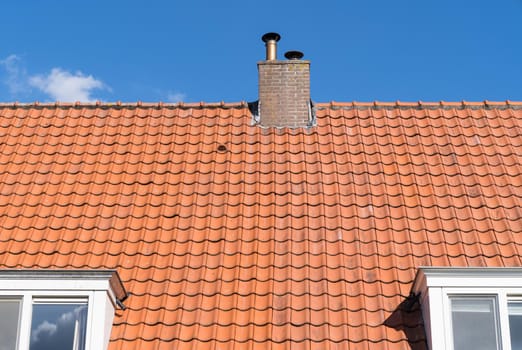Roof with red roof tiles and chimney and a clear blue sky with some clouds on a sunny day