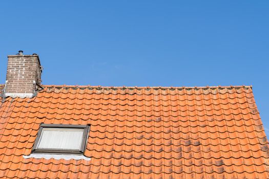 Roof with red roof tiles, a window and chimney and a clear blue sky with some clouds on a sunny day
