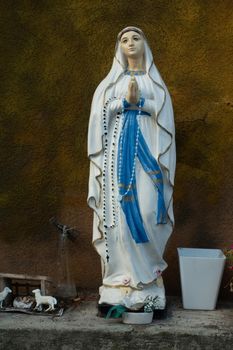 Statue of Virgin Mary as symbol of love and kindness with offerings standing on a stone ledge against a brown wall