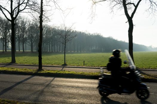 A silhouette of a motor cyclist with trees in the morning sun with mist and a wide view over a grassland