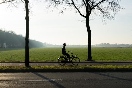 A silhouette of a cyclist looking on his phone with trees in the morning sun with mist and a wide view over a grassland