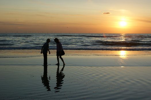 Dark silhouettes of two anonimous children against a sunset sky at the beach with reflection and windmills at the horizon