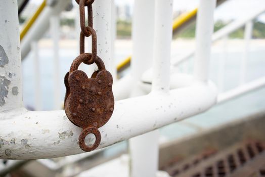 A brown rusty metallic lock hangs on a rusty white lattice of a river embankment railing with a depth of field