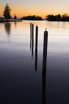 a Row of poles in smooth water on the shore of a lake at dusk showing the reflection of sunset in the water a concept of perspective or the fall of the evening