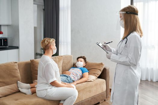 Caring mother sitting on couch near her sick son while competent doctor in medical uniform standing near and writing on clipboard. Pediatrician visiting patient at home.