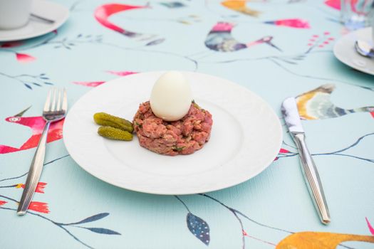 Raw steak tatar with a boiled egg and gurkins on a white plate with cuttlery on a decorative set table