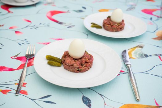 Raw steak tatar with a boiled egg and gurkins on a white plate with cuttlery on a decorative set table