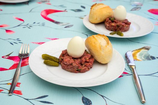 Raw steak tatar with a boiled egg and gurkins on a white plate with cuttlery on a decorative set table