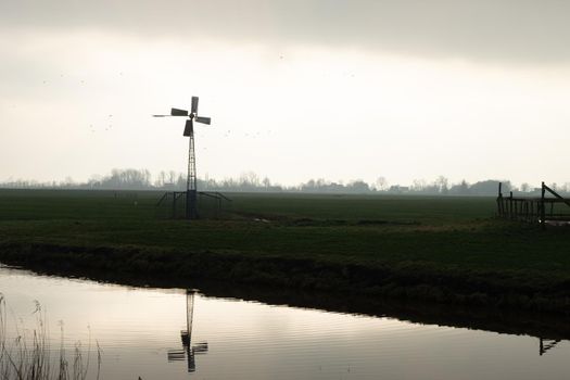 A silhouette of a small windmill in an agricultural setting used to drain the land in wetlands, the polder landscape in the Netherlands at sunset with a river and a cloudy sky