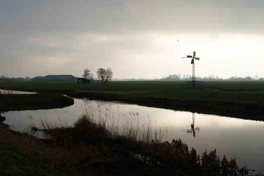 A silhouette of a small windmill in an agricultural setting used to drain the land in wetlands, the polder landscape in the Netherlands at sunset with a river and a cloudy sky