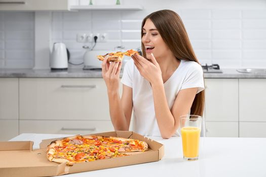 Young brunette with long hair smiling and eating tasty pizza in kitchen. Concept of lunch or dinner at home.