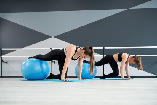 Side view of two attractive fit women training core muscles with one leg on big blue fitness balls in studio with handrails. Gorgeous fitnesswomen practicing plank position on mats. Concept of sport.