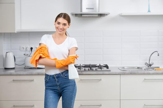 Smiling young woman standing in kitchen with rag and detergent. Concept of cleaning in kitchen.