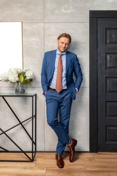 The handsome stylish young man stands at a wall, long curly hair, he is dressed in a expensive blue suit and trousers, he is looking at the camera, a brown classical shoes, Flowers on a curbstone. High quality photo