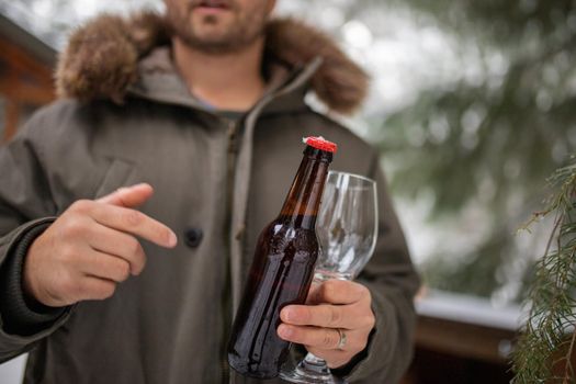 Male hands holding bottle of beer and glass goblet with blurry pines as background. Man preparing to serve beer in the middle of a snowy forest. Relaxing winter holiday