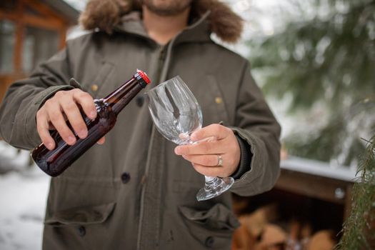 Man pouring beer into glass goblet with cabin and snowy forest as background. Male hands holding bottle and glass in the woods. Relaxing winter holiday