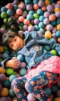 Little asian girl having fun in ball pit with colorful balls. Child playing on indoor playground. Kid jumping in ball pool.