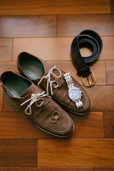Brown mens suede shoes with wristwatch, belt and wedding rings of the bride and groom on a wooden texture. High quality photo