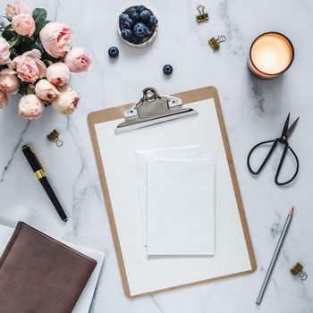 Wedding mock up. Flat lay with blank greeting card, envelope. Clipboard, flowers, scented candle on white marble. Feminine home office mock up with copy space for text or design. Flat lay