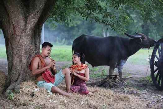 Couple love of Asian Young man and women sitting under tree against buffalo and natural background, rural way of life in the Northeast of Thailand. A young man was blowing a bamboo mouth organ to his lover.