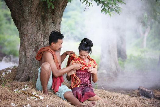 Couple love of Asian Young man and women sitting under tree against buffalo and natural background, rural way of life in the Northeast of Thailand. A young man was blowing a bamboo mouth organ to his lover.