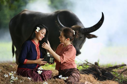 Two Asian woman wearing traditional thai Esan custom style culture sitting in field, while listening  vintage style radio against buffalo and farm background.