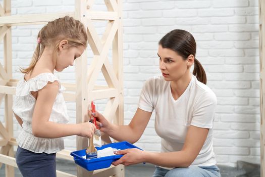 Close up of mother and daughter holding brushes and white paint. Concept of preparing painting rack.