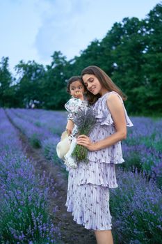 Side view of woman posing with baby girl on hands in beautiful summer lavender field. Young mother wearing dress carrying aromatic bouquet of purple flowers. Concept of nature beauty.
