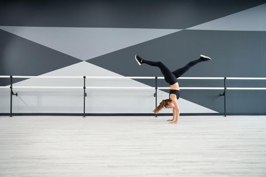 Side view of flexible adult female dancer training in dance hall, handrails on background. Strong fit woman practicing wheel in jump, doing split in air. Concept of choreography, healthy lifestyle.