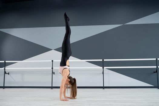 Strong fit woman wearing black tight sportswear standing on arms. Side view of attractive flexible female dancer training before competition in dance hall with ballet handrails. Gymnastics concept.