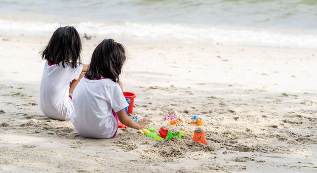 Twin girls while playing beach toys and sitting on a beach sand with white sand and waves