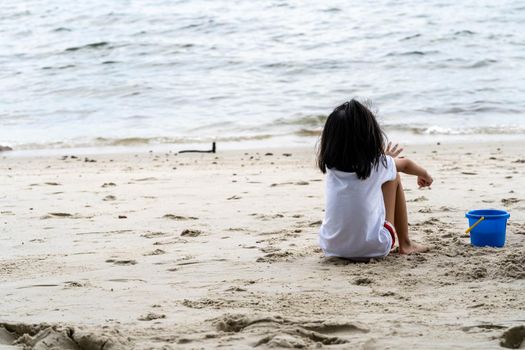 Little girl while sitting on a sand in a beach and looking at the ocean alone