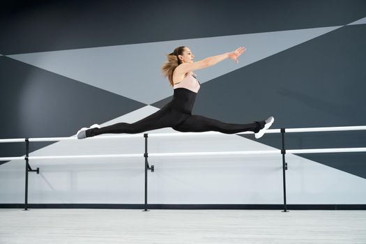 Side view of flexible young female dancer jumping in dance hall with ballet handrails, hi tech interior. Strong fit girl wearing tight sportswear stretching and practising split in air. Sport concept.