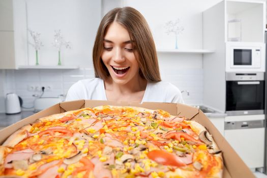 Happy young woman holding hot pizza in box at home. Concept of dinner in kitchen.