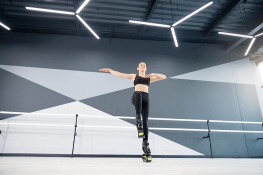 From below view of active female teenager doing cardio workout in hall, hi tech interior. Young pretty girl wearing black sportswear practicing dance moves while doing kangoo jumps and smiling.