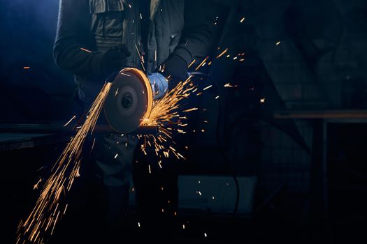 Close up of fitter in special suit cutting with rotating disc for metal with large sparks. Concept of process polishing or welding metal with flash sparks in factory.