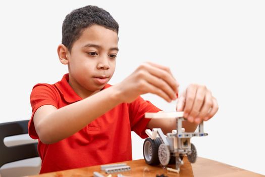 Front view of lovely conentrated african boy sitting at table and using building kit for kids, children creating toys, having positive emotions and joy. Close up of boy working on project.