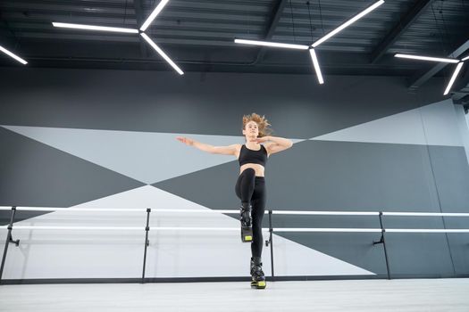 From below view of active female teenager doing dance cardio workout in hall with hi tech interior. Young pretty girl wearing black sportswear practicing dance moves while doing kangoo jumps.